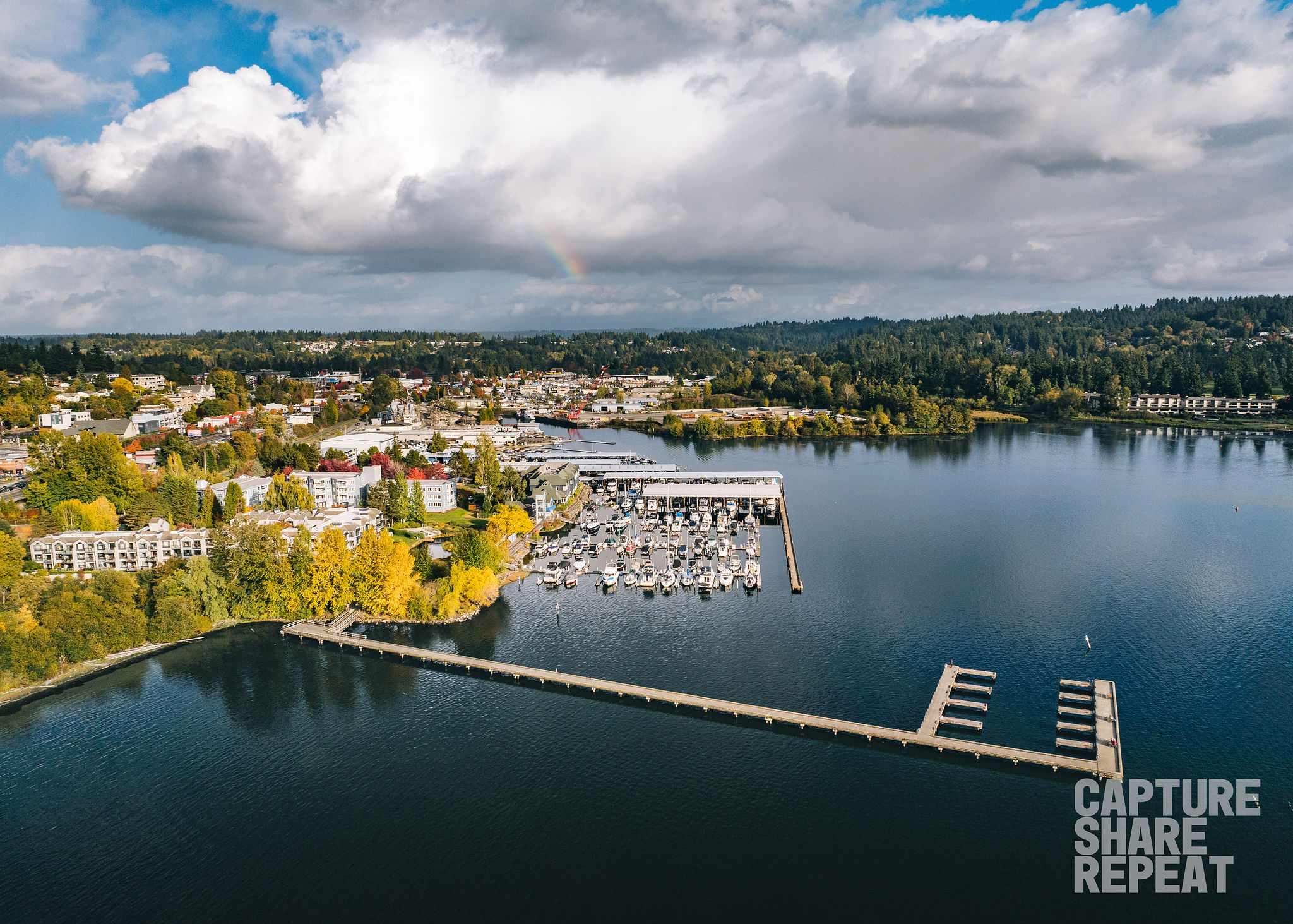 Aerial photo of a lakeside town with a large dock jetting out into the lake. In the middle there is a marina with boats on the lake. The shoreline if filled with trees and the sky is full of white fluffy clouds and a blue sky backdrop and a hint of small rainbow. 